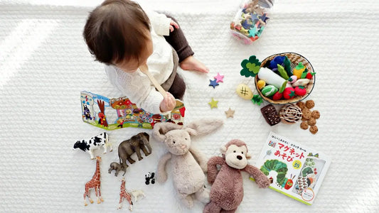 A baby sits on a white blanket surrounded by various toys, including stuffed animals, plastic animals, colorful wooden stars, and picture books.
