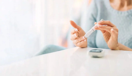 A person with diabetes is holding a blood sugar test strip and pricking their finger. A glucometer lies on the white surface in front of them.