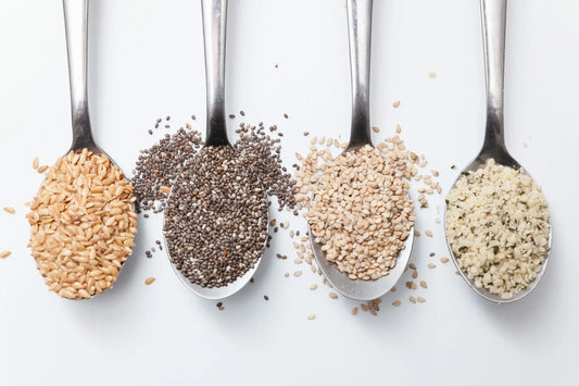Four metal spoons filled with different types of seeds are lined up against a white background.