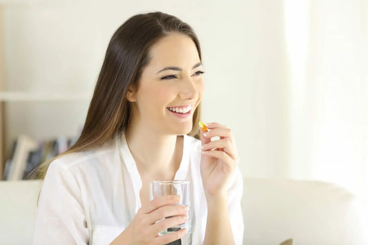 A smiling woman with long brown hair holds a glass of water in one hand and a pill in the other.