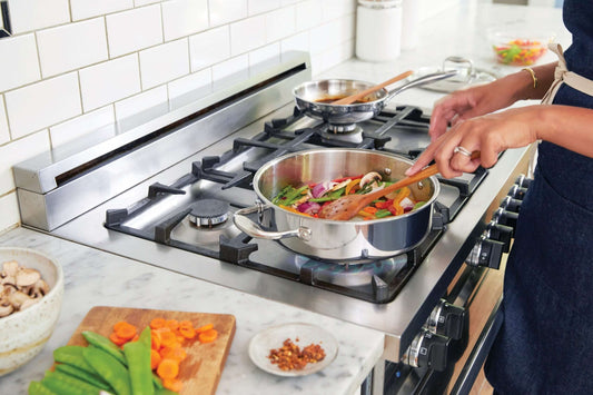 A person wearing an apron cooks vegetables in a stainless steel pan on a gas stove.