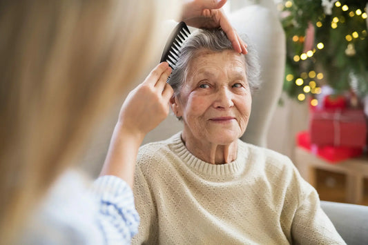 An elderly woman with gray hair and wearing a beige sweater sits in a chair, smiling contentedly as another person combs her hair.
