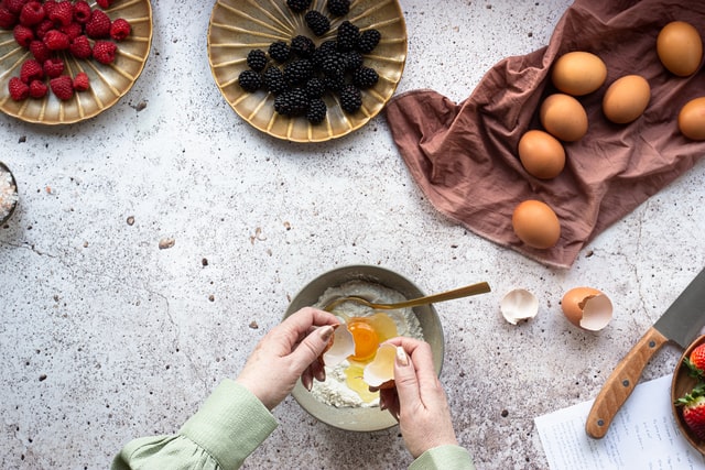 A person in a green shirt is cracking an egg into a bowl with flour on a speckled countertop.