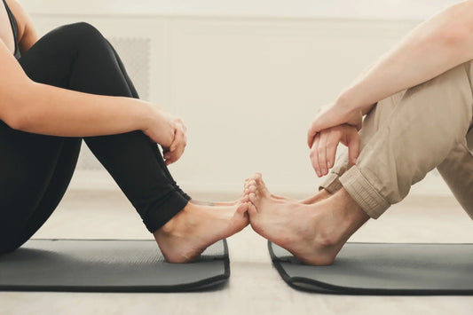 Two individuals sit on yoga mats facing each other, with legs bent and feet touching.