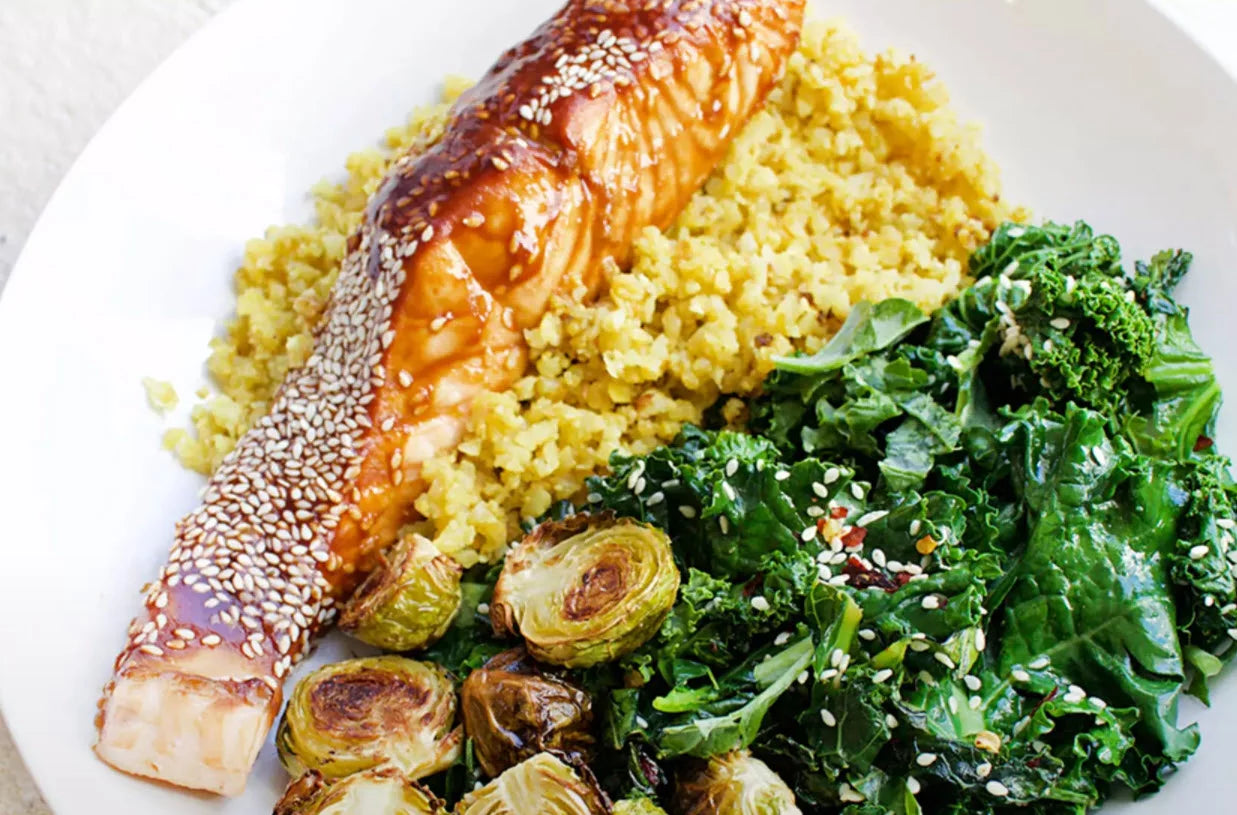 A white plate filled with a sesame-topped salmon fillet, cooked brussels sprouts, a serving of sautéed kale, and a mound of turmeric-seasoned couscous.
