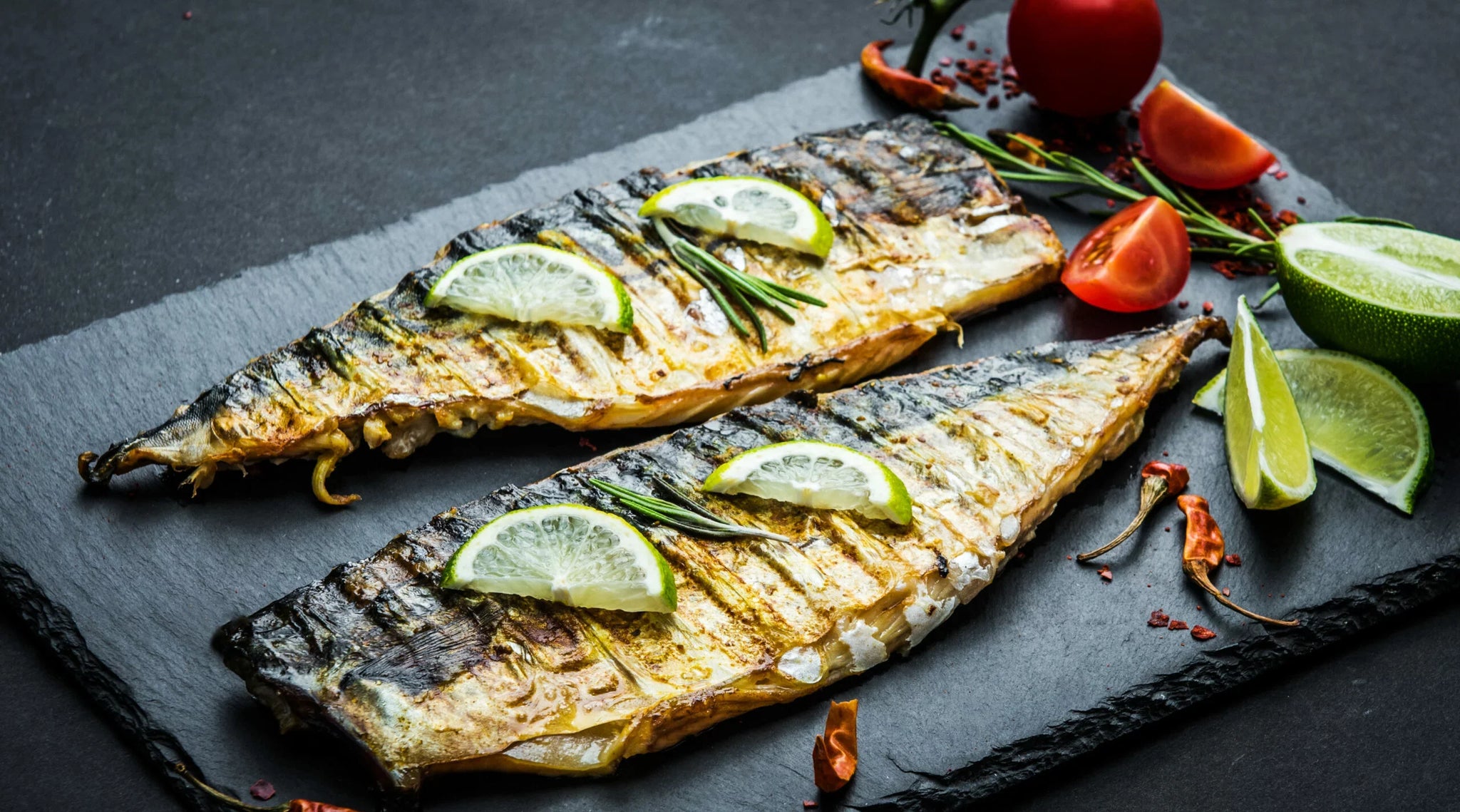Two grilled fish fillets garnished with lime wedges and rosemary sprigs are placed on a dark slate plate.