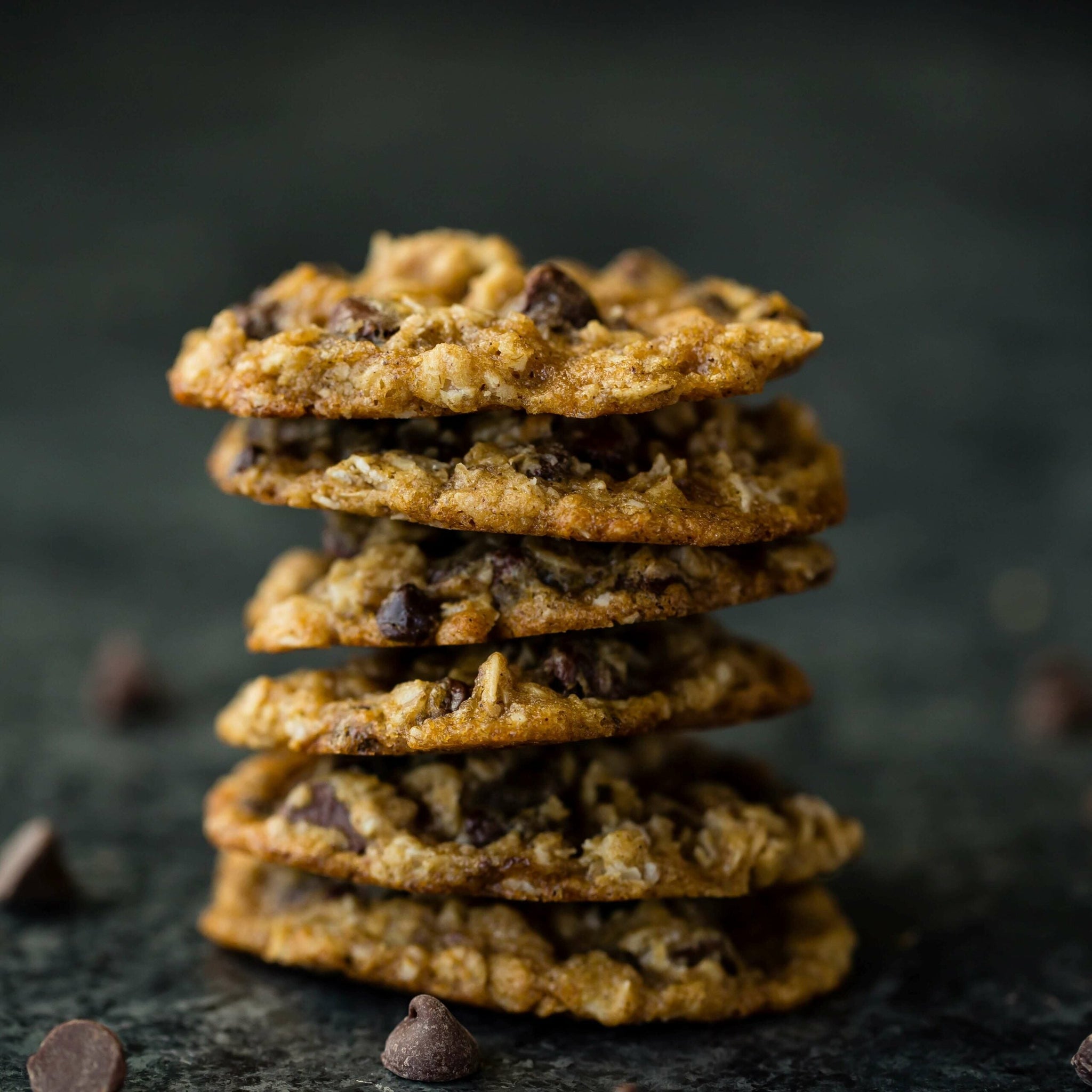 A stack of five chocolate chip oatmeal cookies is placed on a dark, textured surface.