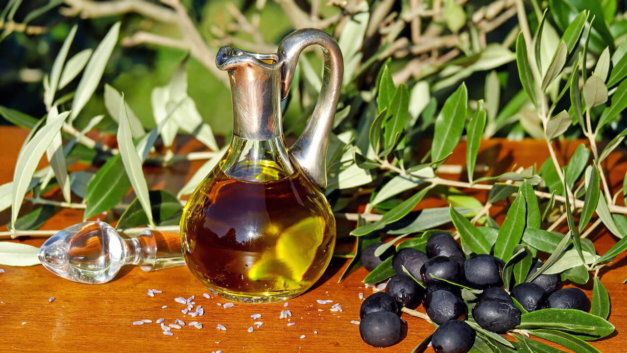 A glass jug filled with olive oil sits on a wooden surface, surrounded by fresh olive branches and a bunch of black olives.