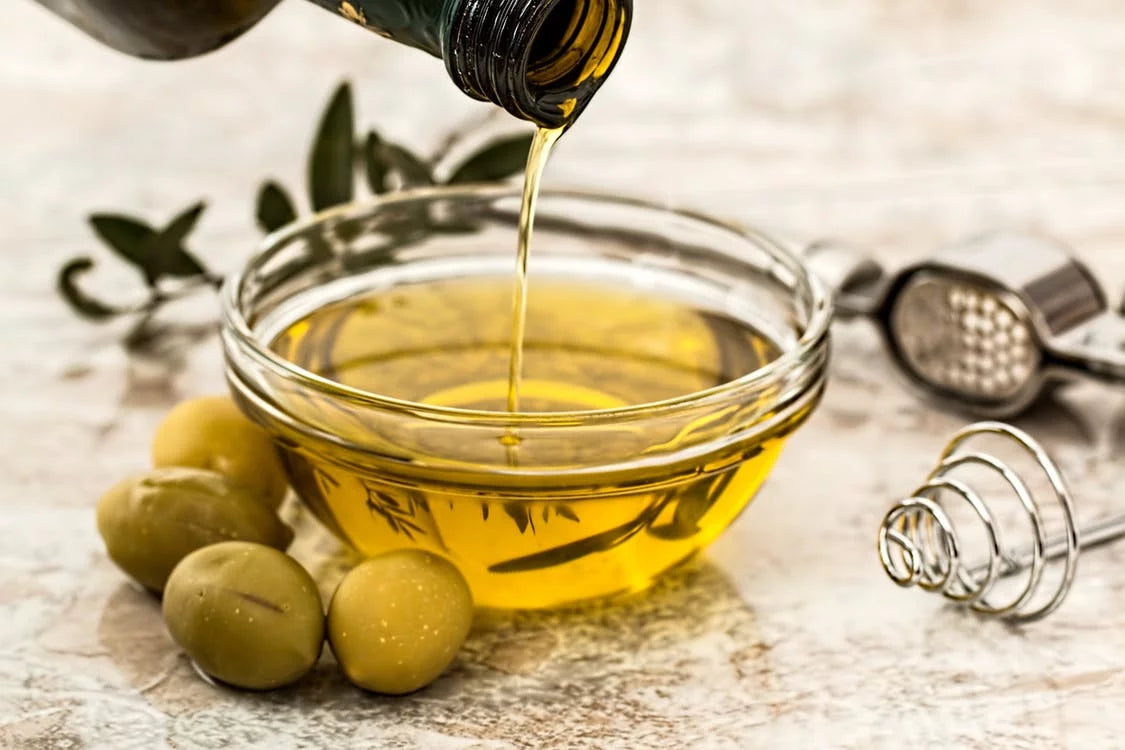 A stream of olive oil is being poured from a bottle into a clear glass bowl filled with glx3 ingredients.