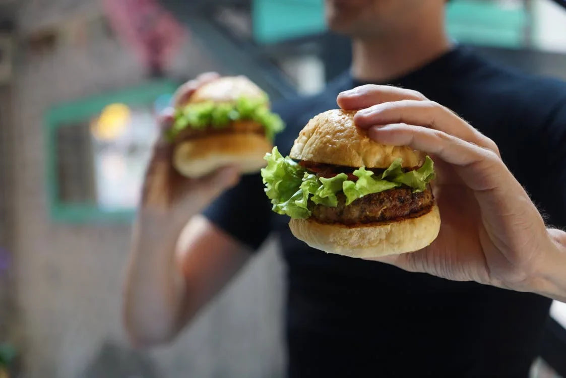 A person in a black shirt holds two burgers, each with lettuce and a patty, in both hands. The background is blurred, focusing on the burgers.