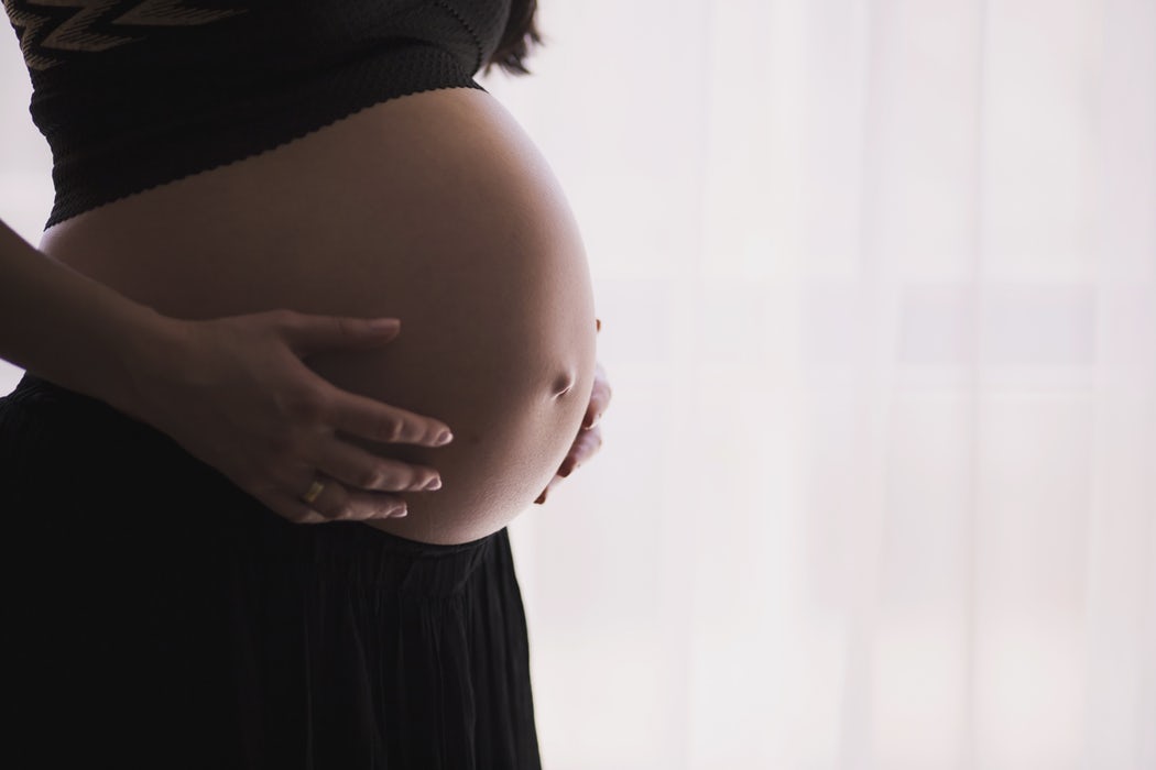 A pregnant woman gently cradles her belly with both hands. She is wearing a black skirt and a dark top.