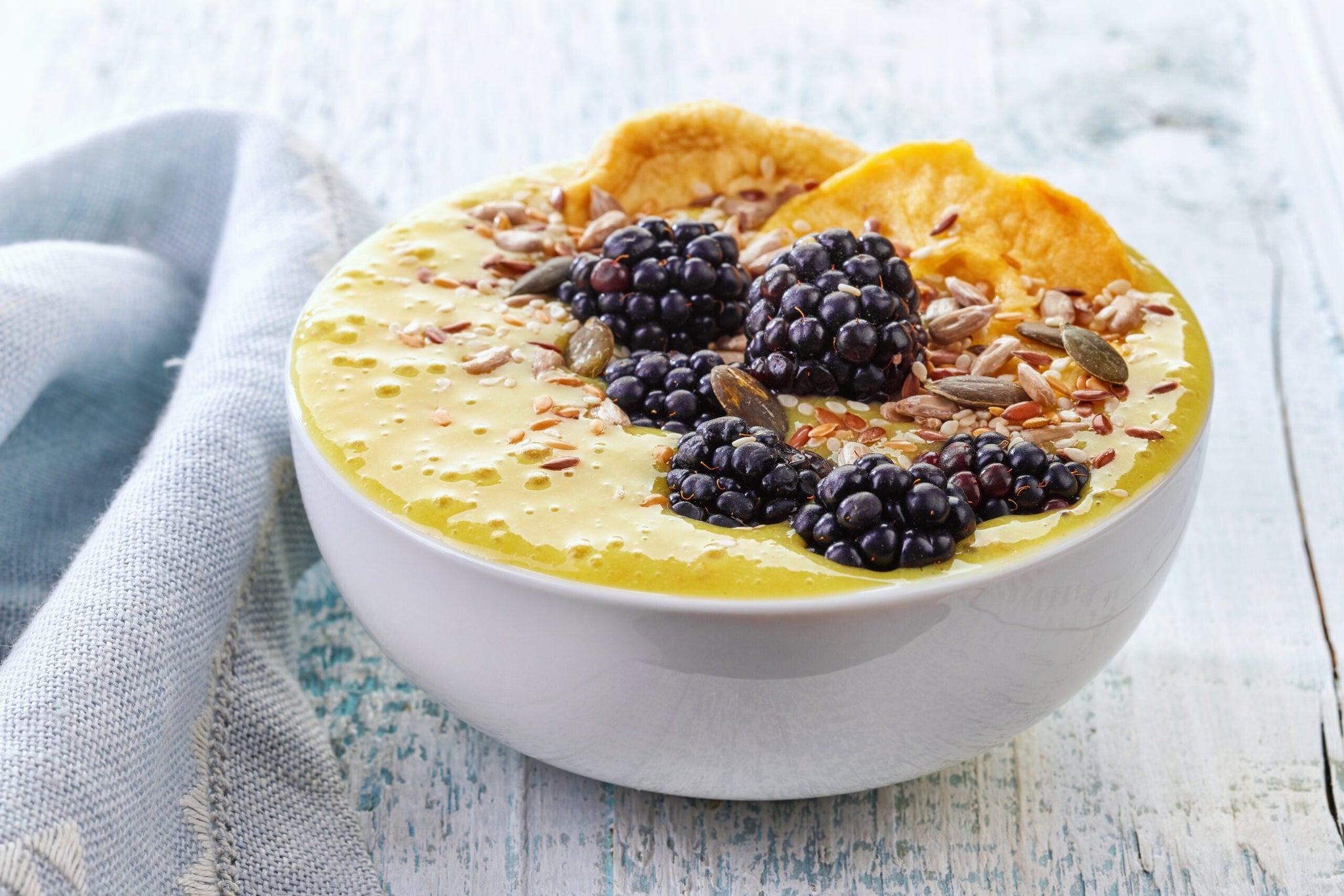 A bowl of creamy yellow smoothie topped with fresh blackberries, dried orange slices, sunflower seeds, and pumpkin seeds.