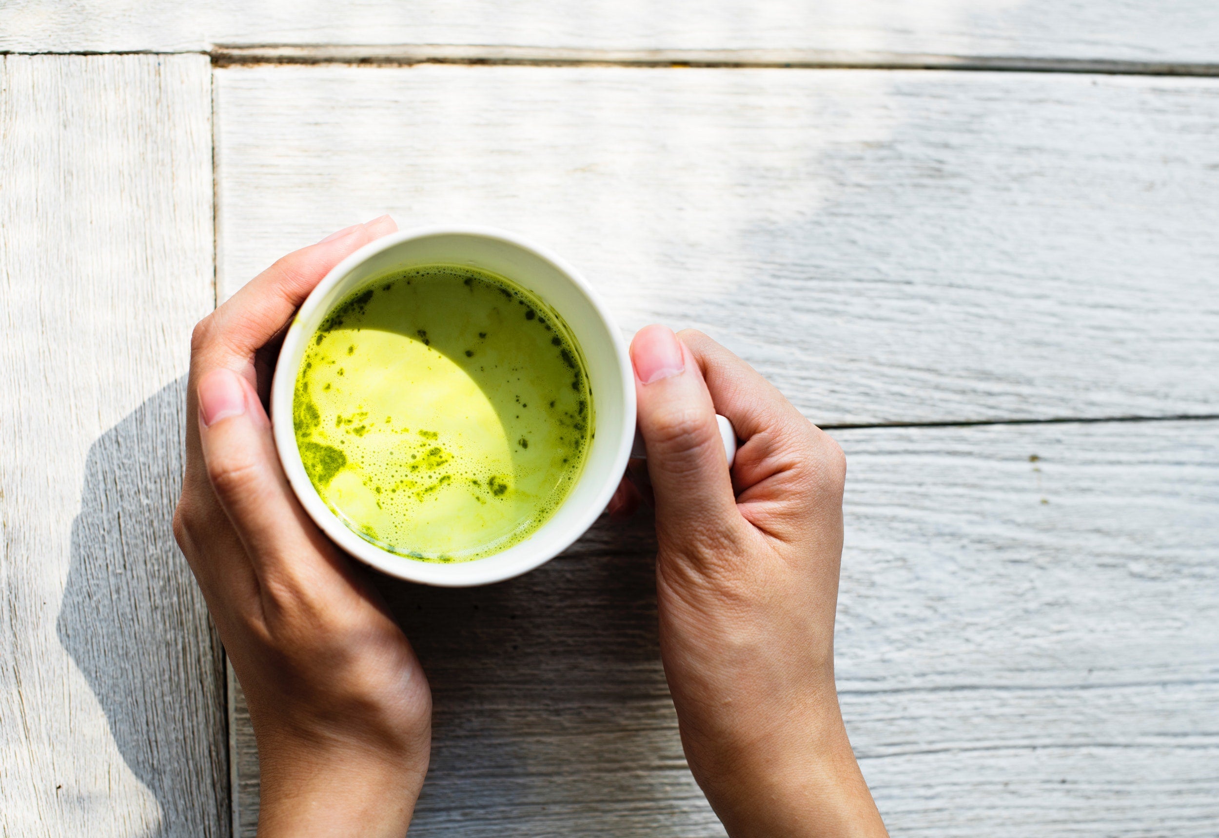 A pair of hands is holding a white cup filled with green tea, one of the finest anti-inflammatory drinks to treat arthritis. The cup is placed on a light-colored wooden surface, and the sunlight casts a soft shadow, creating a serene and calming atmosphere.